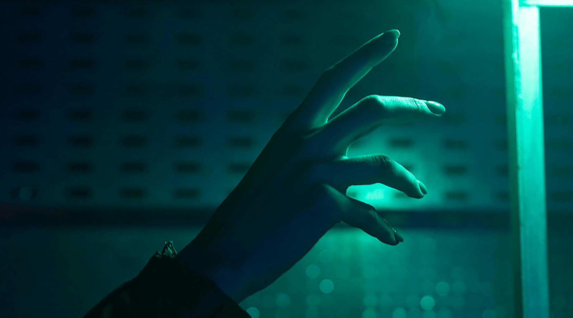 A female hand bathed in blue-green light reaches for a glowing object in the top right hand corner