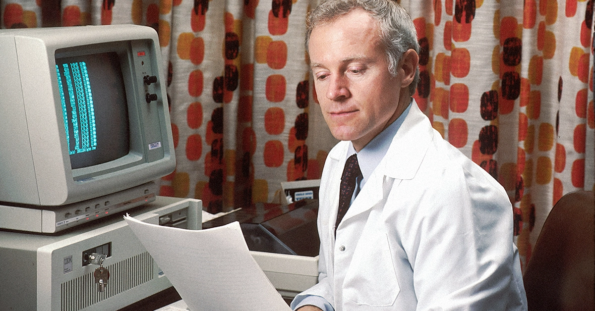 Photo of a man in a white lab coat reading a report in front of a classic computer. Very 80s