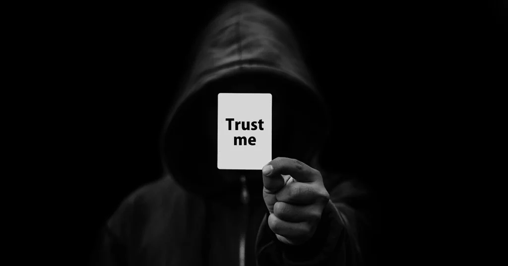 Photo of a man in shadows hiding in a hoodie, hold up a playing card that says "trust me."