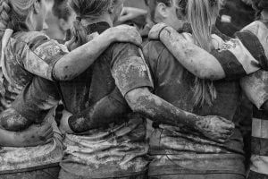 A black and white photograph of a group of female rugby players covered in mud, huddling together.