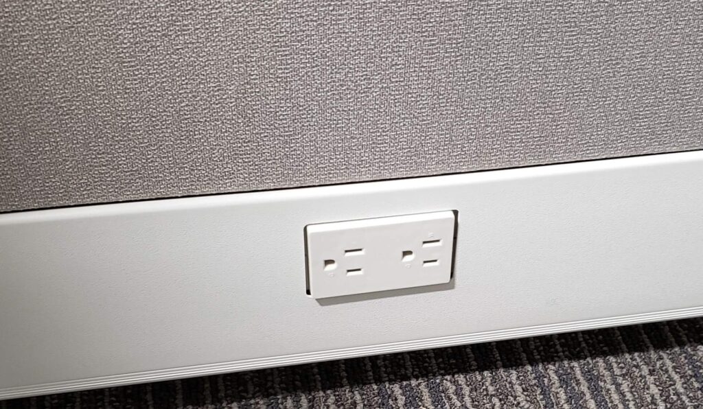 A photo of an electrical outlet in the bottom of an office cubicle wall.