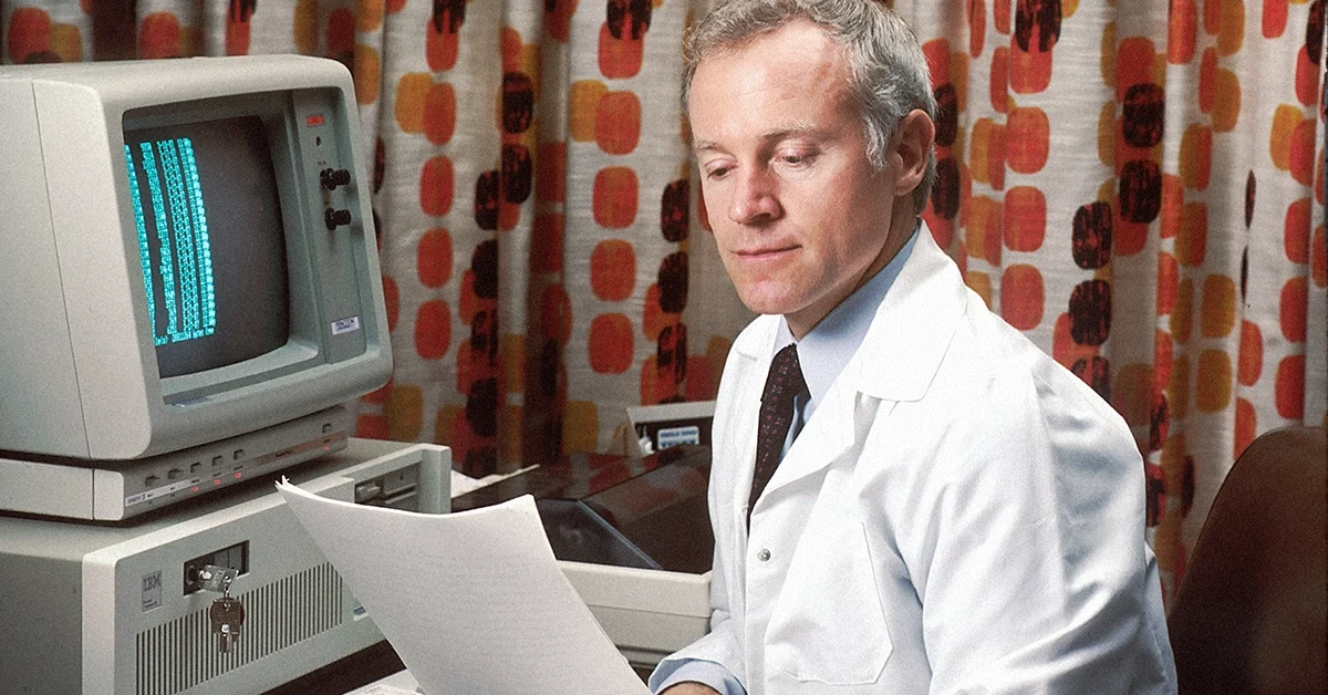 Photo of a man in a white lab coat reading a report in front of a classic computer. Very 80s
