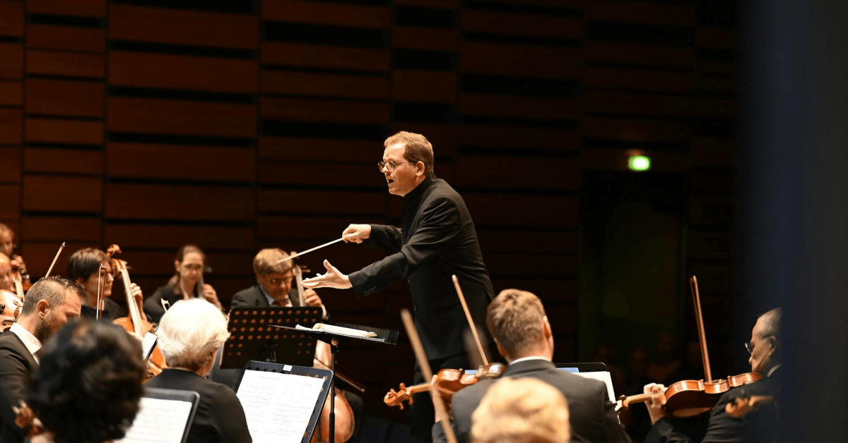 A conductor stands over the orchestra. Photo.