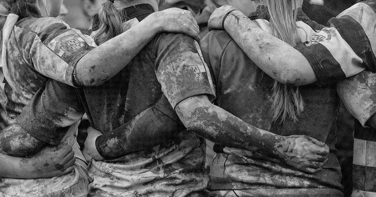 A black and white photograph of a group of female rugby players covered in mud, huddling together.